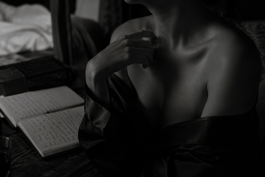 Black and white fine art photograph of a partially draped figure writing in a journal, featuring soft low-key lighting, an intimate atmosphere, and elegant sensual portrait photography